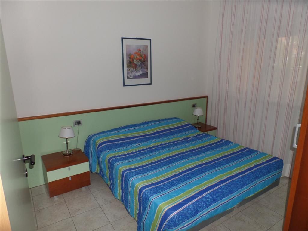 A bedroom with a bed, two bedside tables, a nightstand, a lamp on each table, a framed picture on the wall, and a window with curtains.