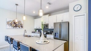 Una cocina moderna con una isla central, electrodomésticos de acero inoxidable, iluminación colgante y un reloj en la pared.