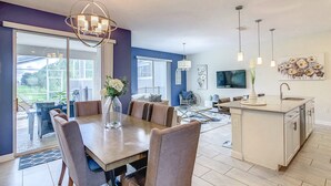 Una cocina moderna con una isla central, una zona de comedor con una mesa grande y sillas, y una sala de estar con sofá y televisor.