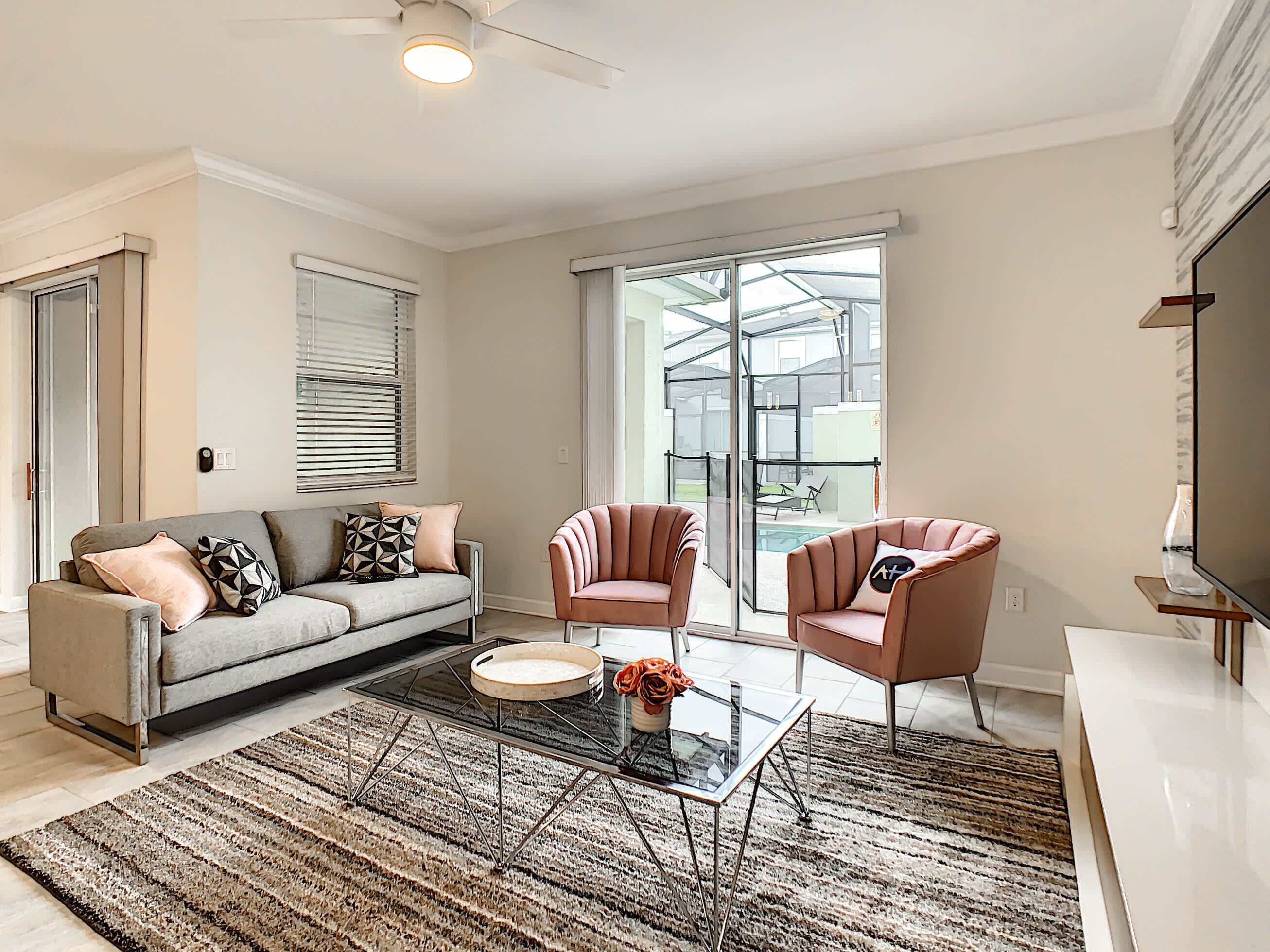 Ein modernes Wohnzimmer mit einem grauen Sofa, zwei Sesseln, einem Glastisch und einer großen Glasschiebetür, die auf einen Balkon führt.