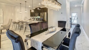 Un comedor moderno con una mesa negra, mantel blanco y sillas oscuras. Un candelabro cuelga encima, y hay lámparas colgantes. Se ve una cocina al fondo con armarios oscuros y electrodomésticos de acero inoxidable.