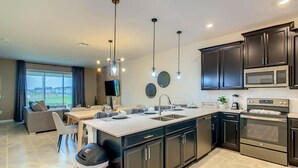 A modern kitchen with dark cabinetry, a central island, and a dining area with a view. - Last Minute Disney Getaway! Themed Bedrooms & Private Balcony. Book Now! #87993 (Orlando)