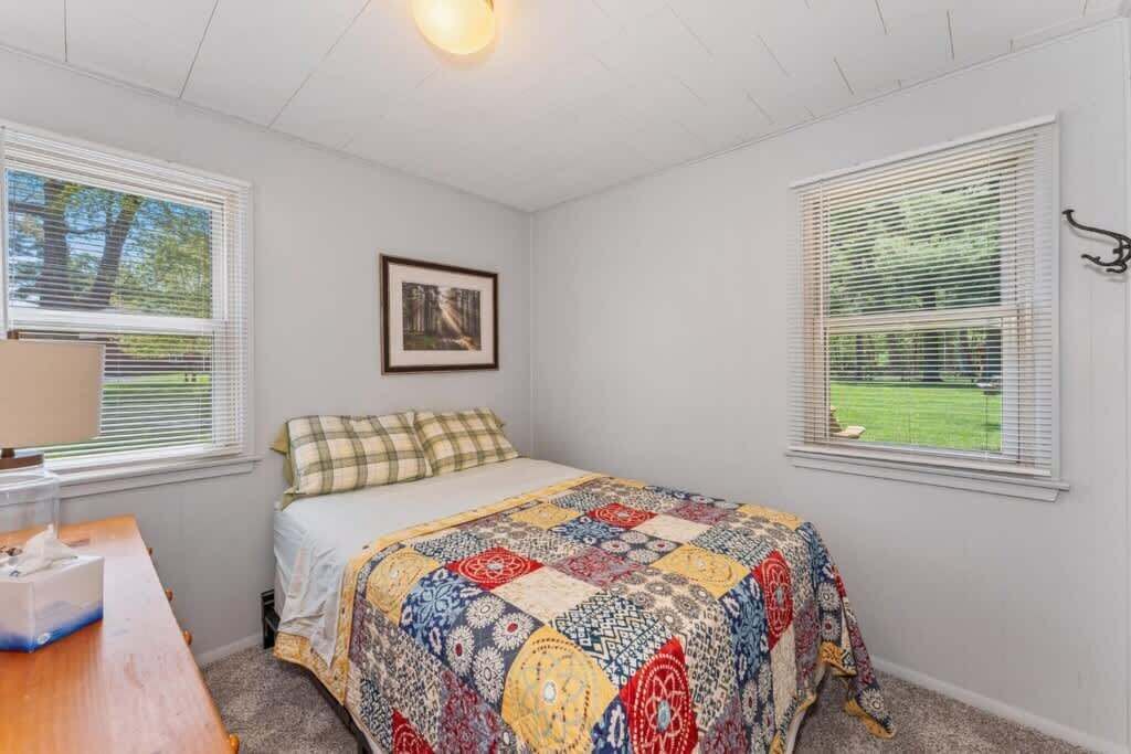 A bedroom with a bed, a bedside table, and two windows with blinds.
