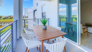 A balcony with a wooden table and white chairs, a potted plant, and a view of a modern building and landscape. - Last Minute Disney Getaway! Luxury Condo with Private Balcony. Book Now! #88039 (Orlando)
