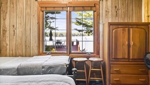 Una habitación con paredes de madera, un ventanal grande, una cama, dos taburetes y un armario.