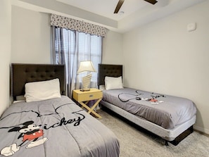 Un dormitorio con dos camas, cada una con cubrecama con temática de Mickey Mouse, una lámpara de noche y una ventana con cortinas.