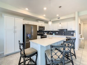 Una cocina moderna con armarios blancos, una isla grande con taburetes de bar, electrodomésticos de acero inoxidable e iluminación colgante.