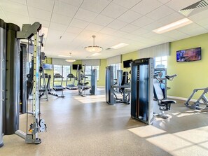 A gym with various exercise machines, a television on the wall, and a window providing natural light.