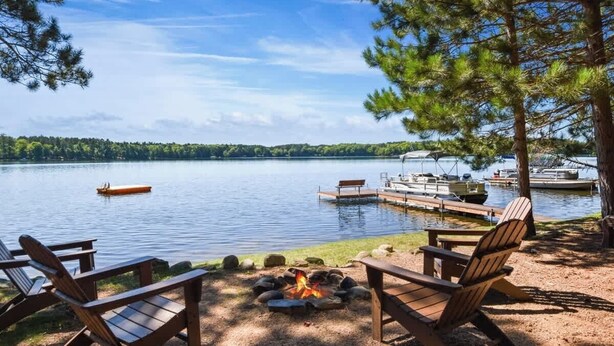 Entorno junto al lago con un brasero, sillas de madera y un muelle con barcos.