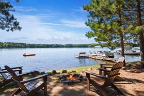 A lakeside setting with a fire pit, wooden chairs, and a dock with boats.