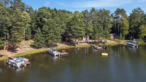 Una propiedad frente al lago con barcos amarrados, una cabaña pequeña y una casa rodeada de árboles frondosos.