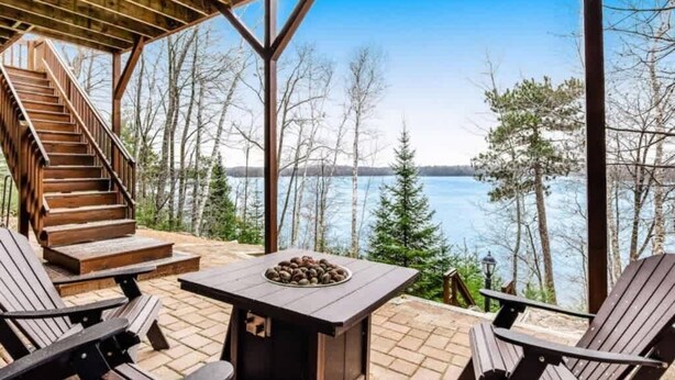 Een overdekt terras met een houten tafel en stoelen, een trap en uitzicht op een meer omgeven door bomen.