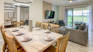 Ein Essbereich mit einem Marmortisch, gedeckt für sechs Personen, Holzsesseln, einem Deckenventilator, einem Fernseher und Blick auf eine grüne Landschaft durch große Fenster.