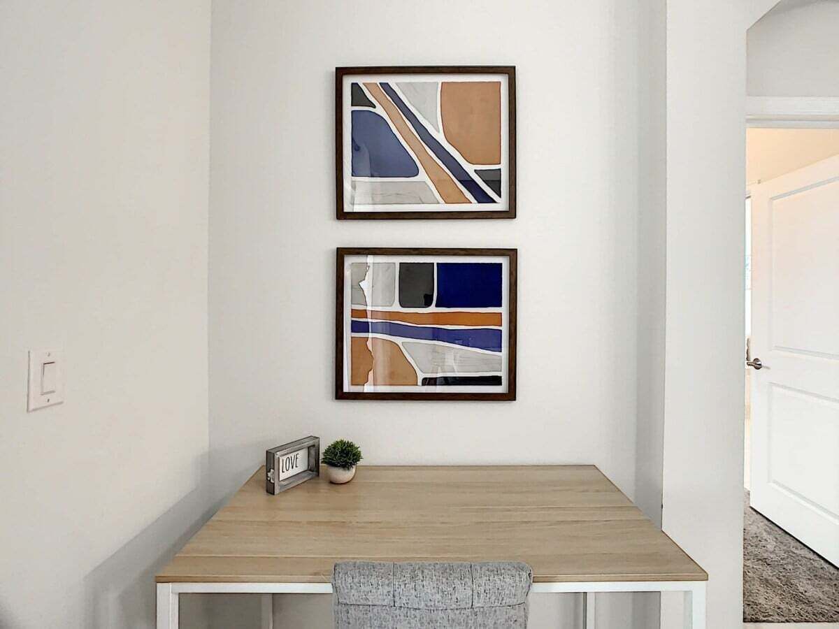 A small, neatly organized room with a wooden desk, two framed abstract paintings, a small potted plant, and a framed sign with the word "LOVE".