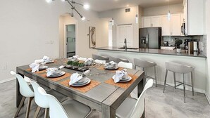 A modern dining area with a large table set for six, a kitchen in the background, and pendant lighting.