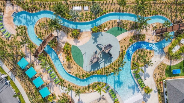 Vista aérea de un complejo turístico con una piscina serpenteante, tumbonas y un pequeño puente.