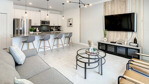 Una cocina moderna con electrodomésticos de acero inoxidable, una zona de comedor con taburetes de bar, una sala de estar con sofá y una mesa de centro, y un televisor montado en la pared.
