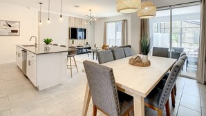 Una cocina moderna con una isla central, zona de comedor con una mesa grande y sillas, y un televisor montado en la pared.