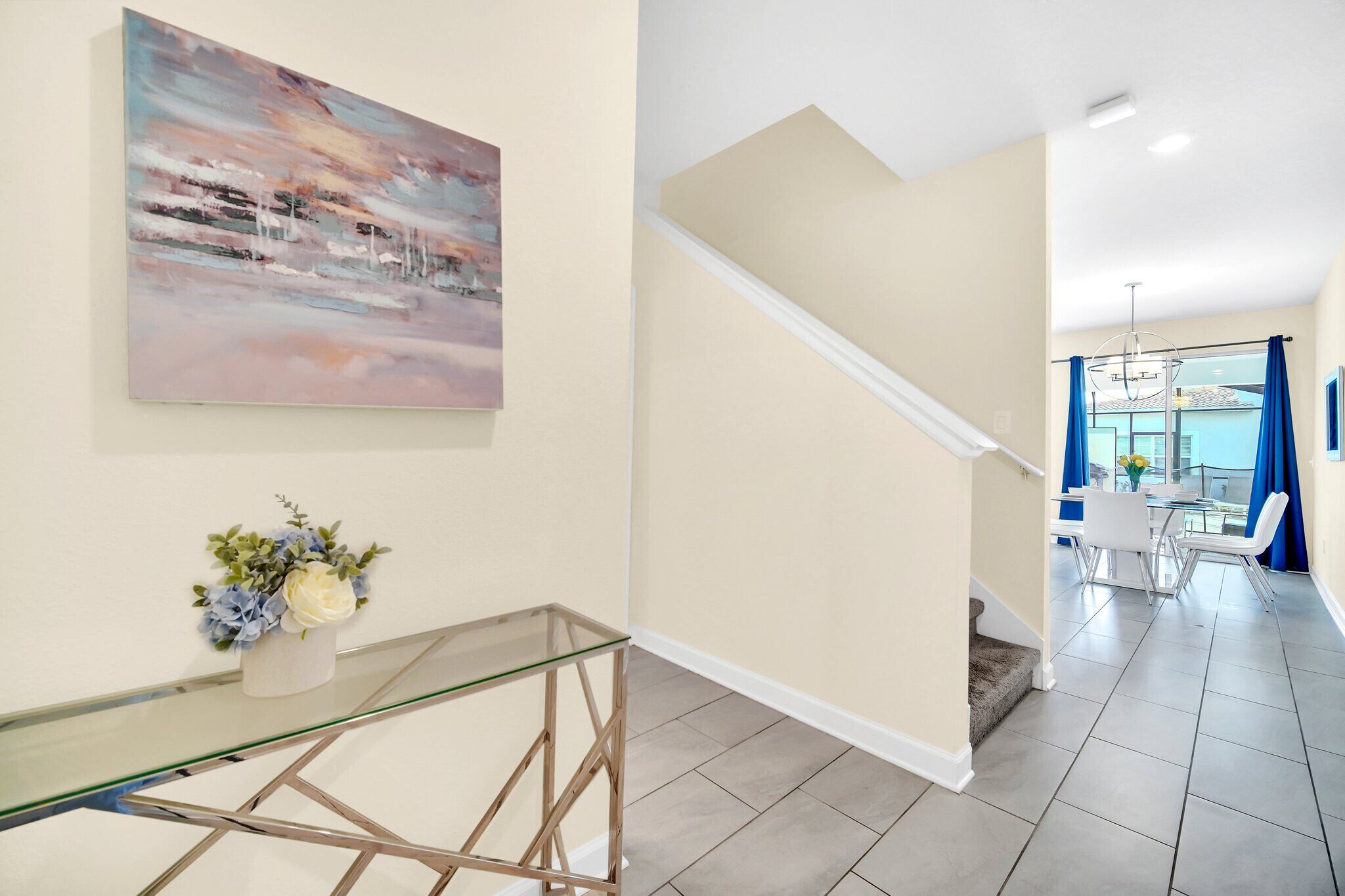 A modern interior with a glass table, a painting, and a staircase leading to a dining area with blue curtains.