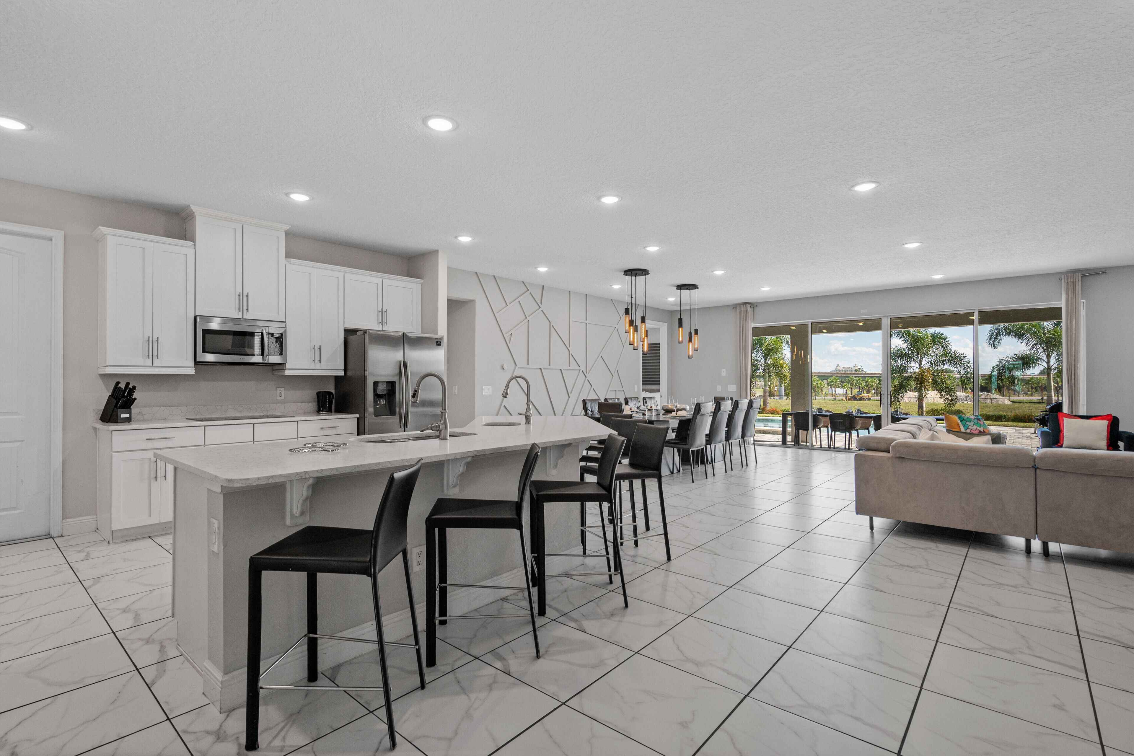 A modern kitchen with white cabinetry, a central island with bar stools, and a dining area with a large window offering a view of the outdoors.