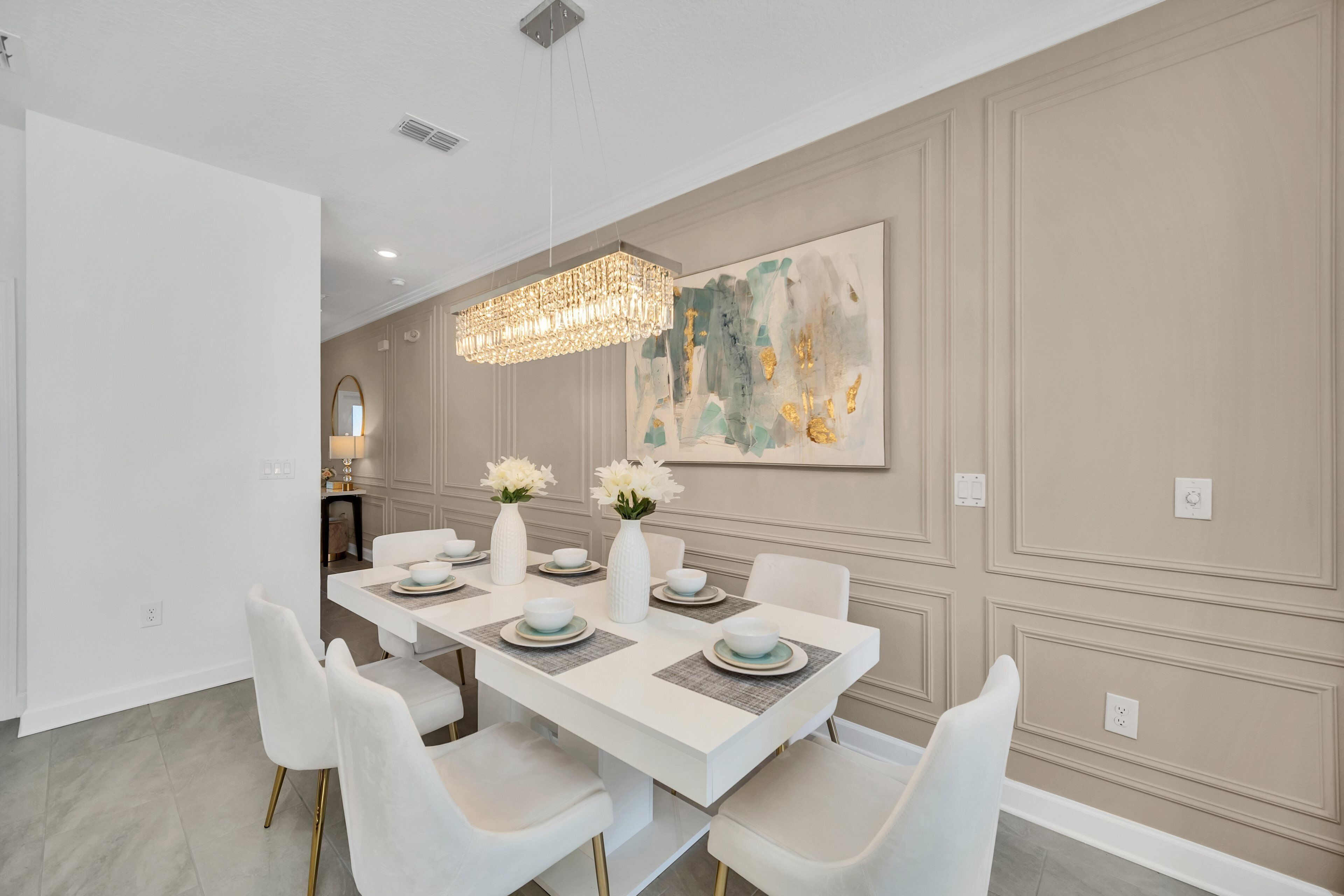 A dining room with a white table set for six, white chairs, a chandelier, and a large abstract painting on the wall.