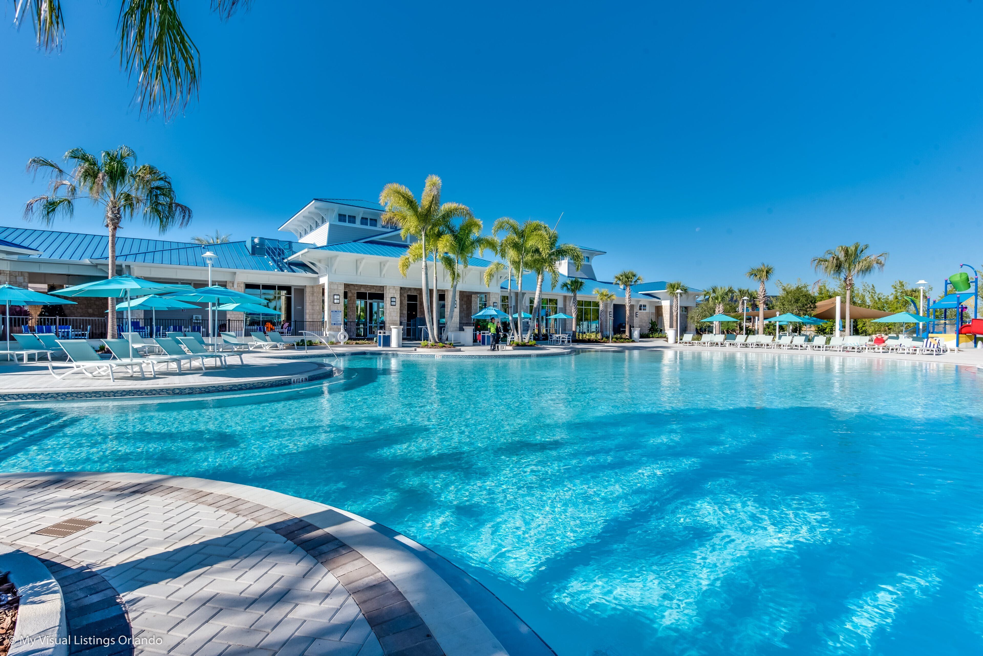 Une grande piscine entourée de chaises longues et de parasols, bordée de palmiers et d’un bâtiment moderne doté d’un toit bleu.