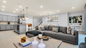 A modern living room with a gray sectional sofa, a coffee table, and a kitchen area with gray cabinets and stainless steel appliances. - Last Minute Disney! Private Pool, Hot Tub, Game Room & Themed Bedrooms. Book Now! #59349 (Orlando)