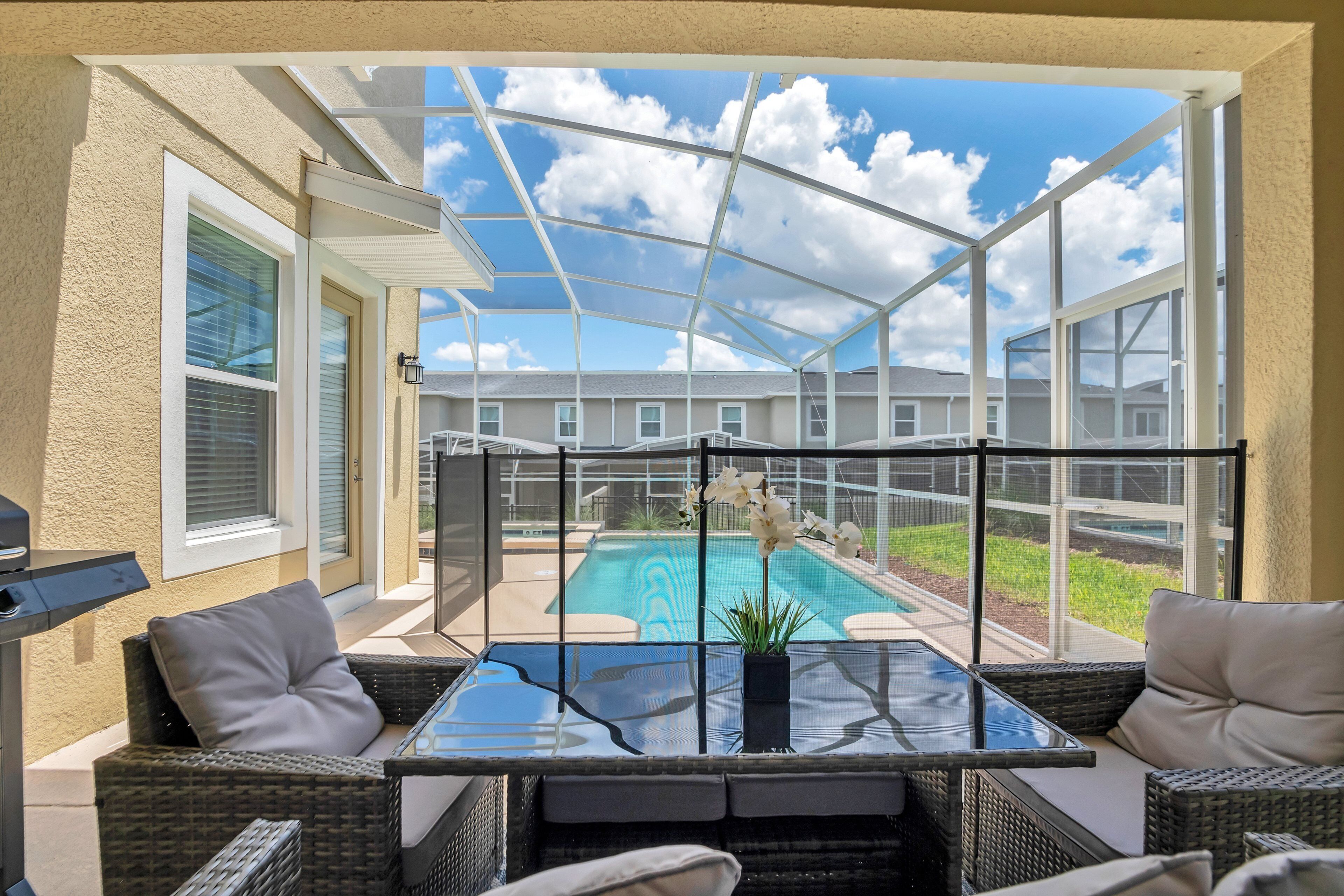 Un patio cubierto con mobiliario de mimbre, una mesa de vidrio y vistas a una piscina.