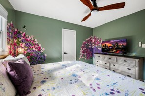 Un dormitorio con cama, cómoda, ventilador de techo, decoración floral en la pared y televisor con imágenes de la ciudad.