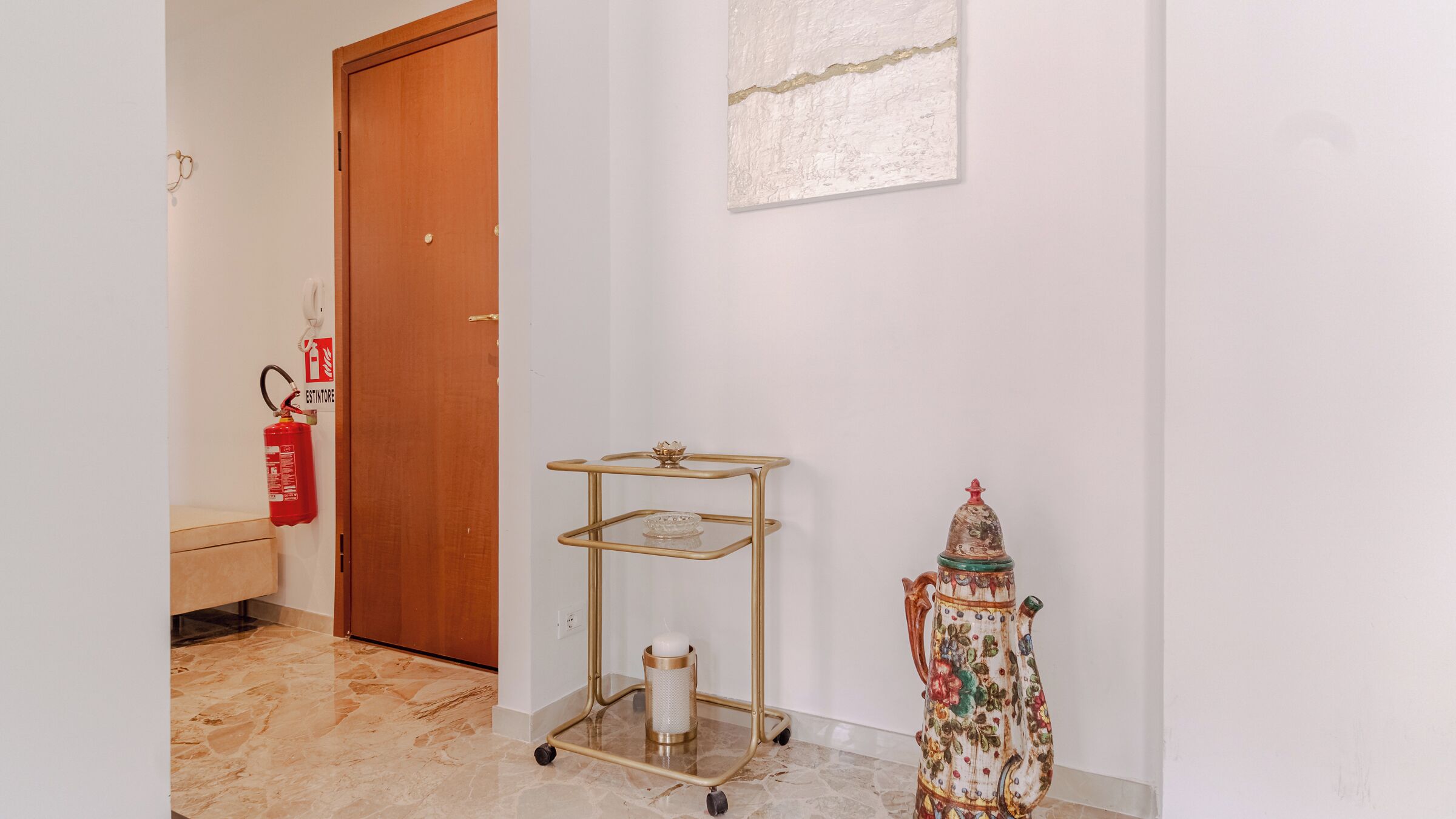 Un corridoio con una porta in legno, un estintore, un carrello metallico e un vaso decorativo.