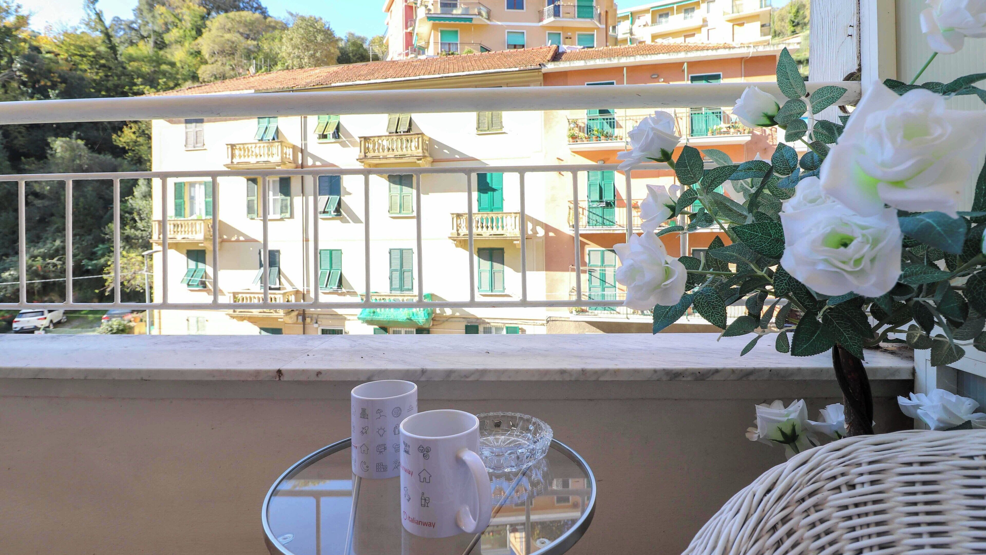 Ein Balkon mit Blick auf ein Gebäude mit Balkonen und grünen Rollläden, ein Korbstuhl, ein kleiner Tisch mit einer Tasse und einer Glasbowl.