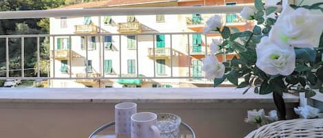 Un balcon donnant sur un immeuble doté de balcons et de volets verts, une chaise en osier, une petite table avec une tasse et un saladier en verre.