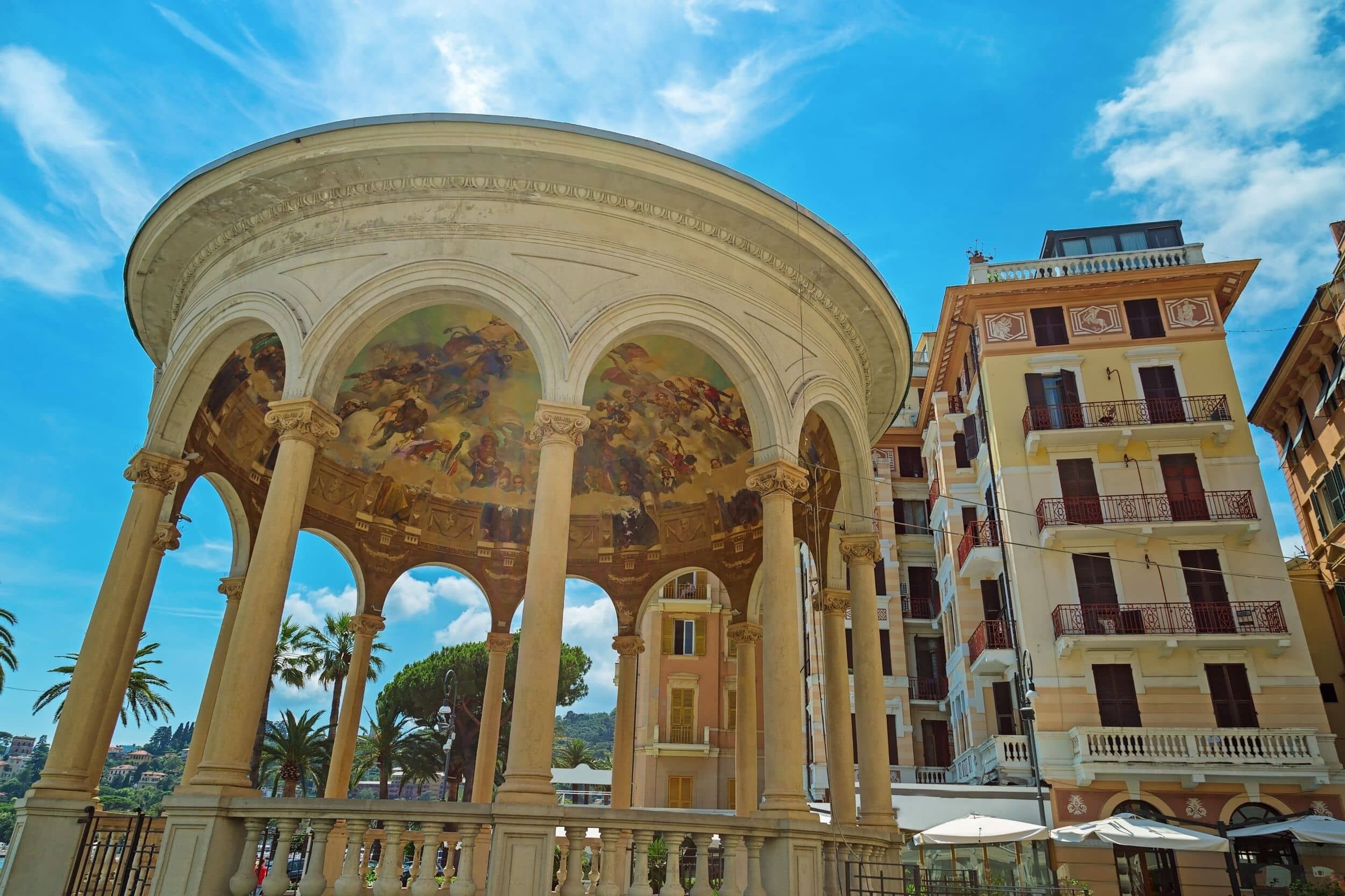 A classical pavilion with ornate columns and a detailed frescoed ceiling, surrounded by urban buildings and palm trees.