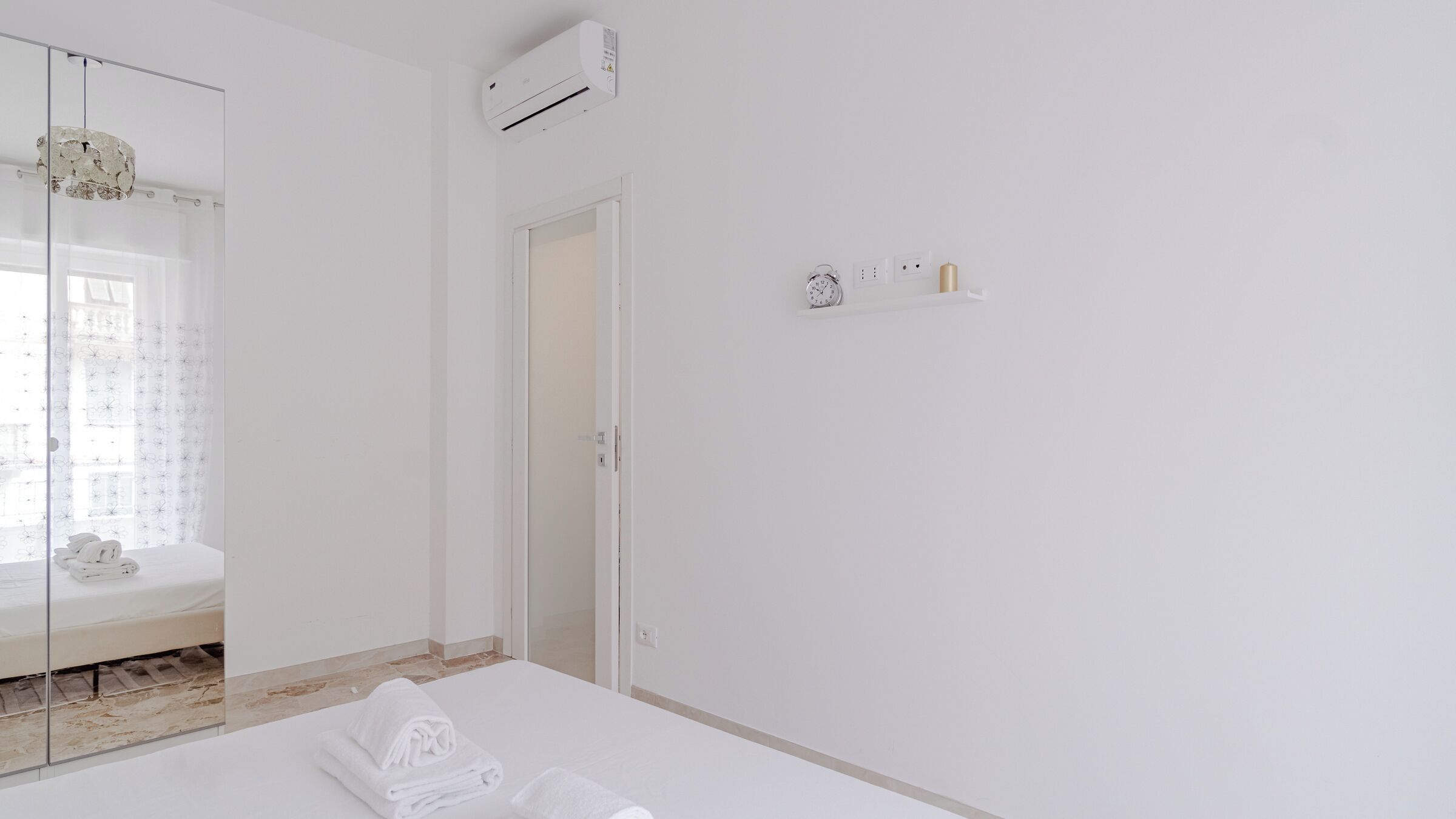 A minimalist hotel room with a bed, a glass shower enclosure, and a wall-mounted air conditioner.