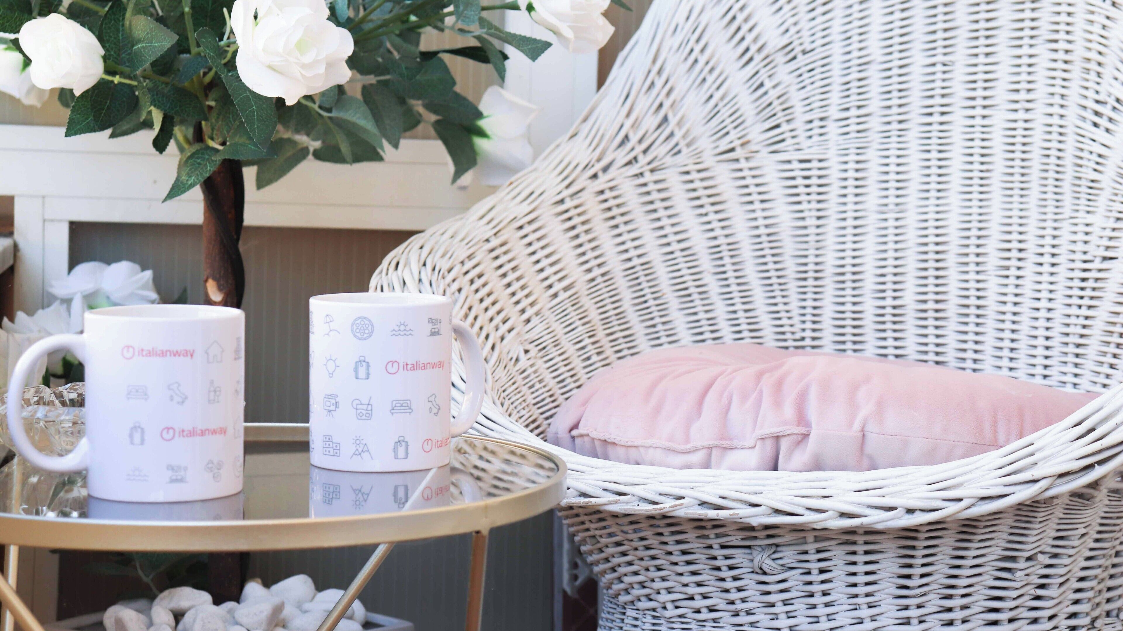 Une chaise en osier avec un coussin rose, une tasse blanche ornée de motifs de voyage et une fleur blanche en arrière-plan.