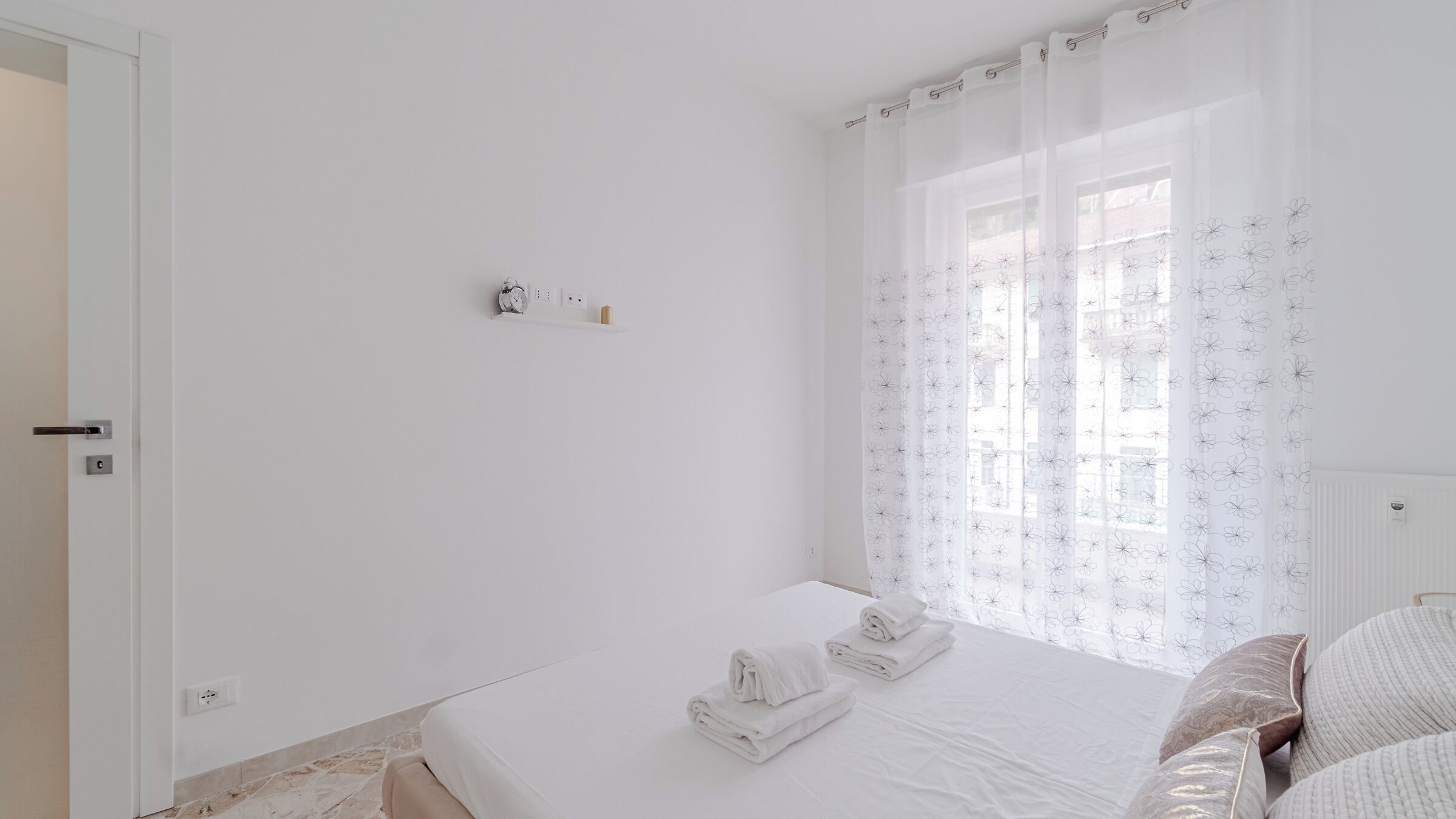 A minimalist bedroom with a bed, a bedside table, and a window with floral curtains.