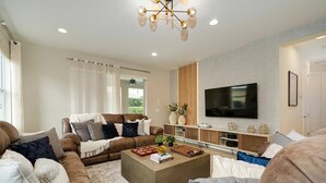 Un salon moderne avec un canapé d’angle, une table basse avec des livres, un téléviseur à écran plat fixé au mur et un luminaire décoratif.