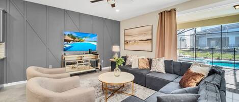 Una sala de estar moderna con un sofá gris, sillones beige, una mesa de centro, un televisor y un ventilador de techo.