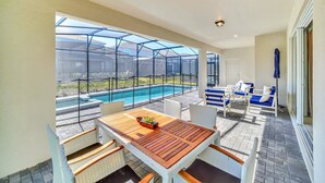 A patio area with a wooden table and white wicker chairs, overlooking a swimming pool with a glass enclosure. - Last Minute Disney! Private Pool, Hot Tub, Game Room & Themed Bedrooms. Book Now! #78231 (Orlando)