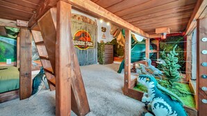 A room with a wooden ceiling, a bed with a green pillow, a dinosaur-themed play area, and a sign reading "Jurassic Park".