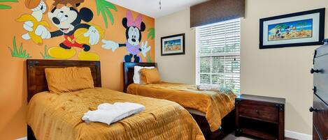 Habitación de hotel con dos camas, ventilador de techo, ventana con persianas y una pared decorada con motivos de Disney.