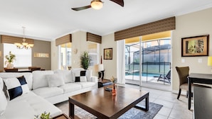 A modern living room with a white sectional sofa, a dark wooden coffee table, and a view of a pool through large sliding glass doors. - Last Minute Disney Getaway! Private Pool, Hot Tub & Game Room. Book Now! #82571 (Orlando)