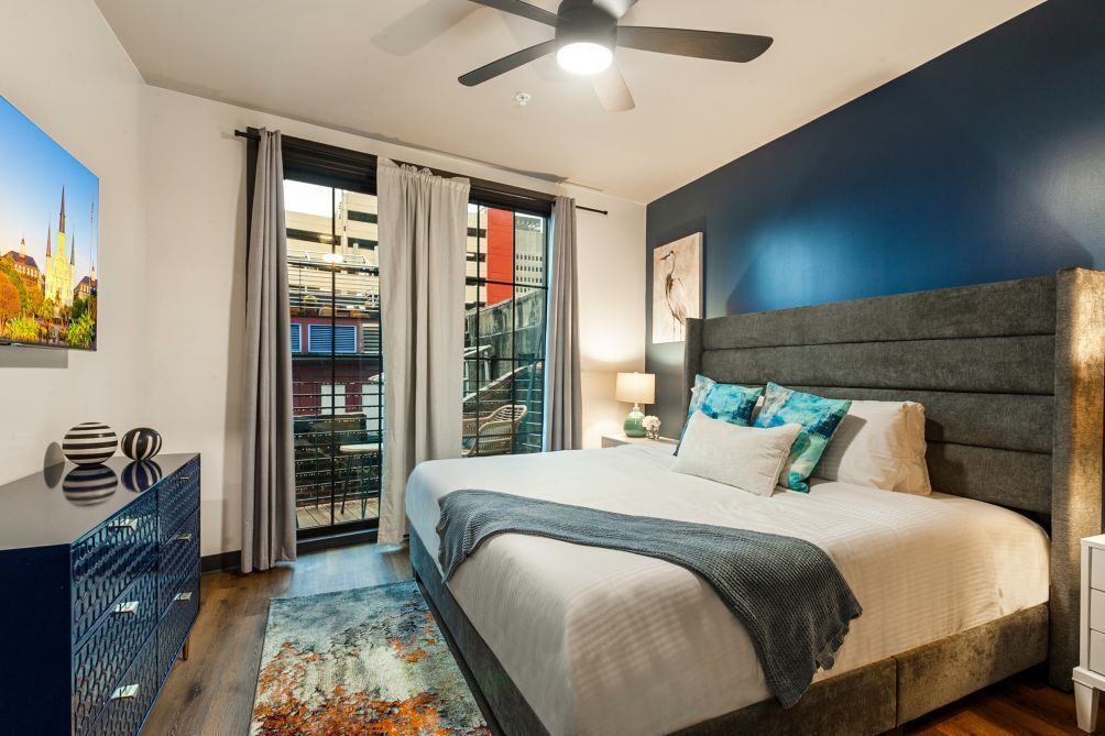 A modern bedroom with a large bed, a ceiling fan, a view of a building through a glass door, and a painting on the wall.