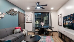 Ein modernes Wohnzimmer mit grauem Sofa, einem runden Couchtisch und einem Esstisch mit Stühlen. Es gibt einen Deckenventilator, ein großes Fenster mit Vorhängen und Bilder an den Wänden.