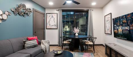 Una sala de estar moderna con un sofá gris, una mesa redonda de centro y una mesa de comedor con sillas. Hay un ventilador de techo, un ventanal con cortinas y obras de arte en las paredes.