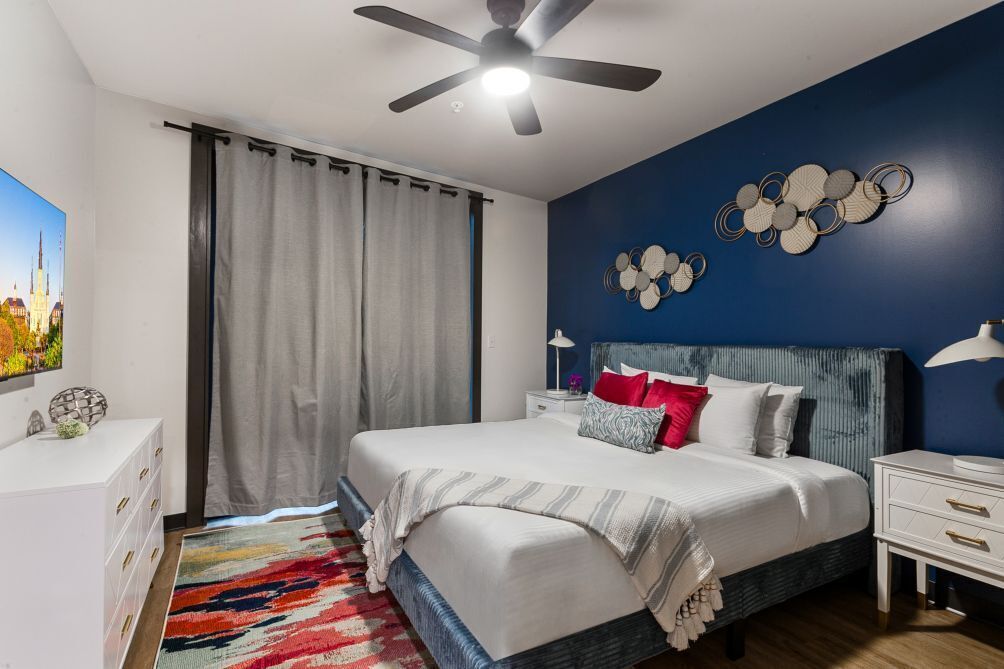 A bedroom with a bed, a ceiling fan, a nightstand, a lamp, and a wall decoration.