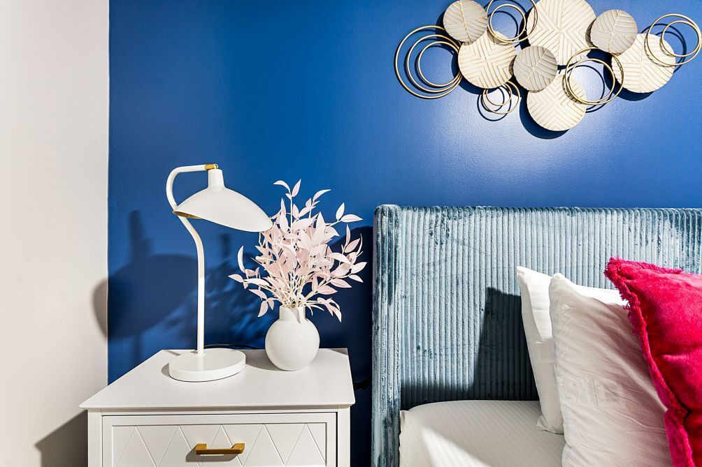 Ein Schlafzimmer mit blauer Wand, einem weißen Nachttisch mit Lampe und Vase, einem blauen Kopfteil und einem roten Zierkissen.