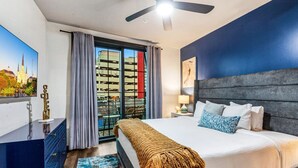 Une chambre d’hôtel avec un lit, un ventilateur de plafond, une table de chevet, une lampe, une grande fenêtre avec des rideaux et un tableau représentant un paysage urbain.