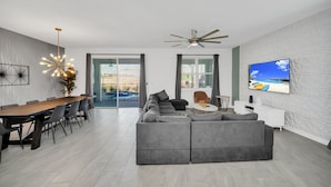 Uma sala de estar moderna com sofá seccional, mesa de jantar com cadeiras, ventilador de teto e televisão na parede.