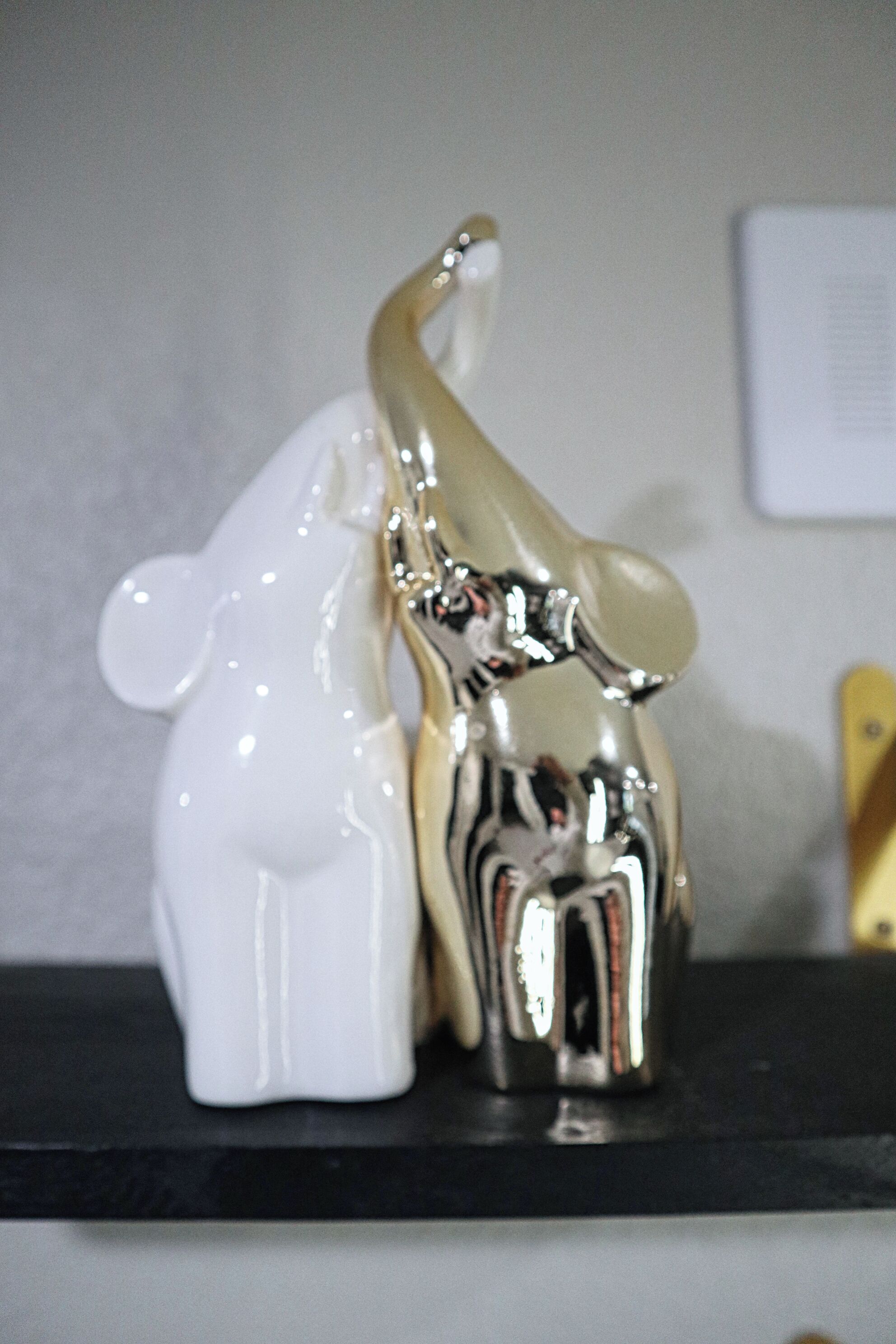 Een witte keramieken olifantje naast een metalen olifantje op een zwart schap.